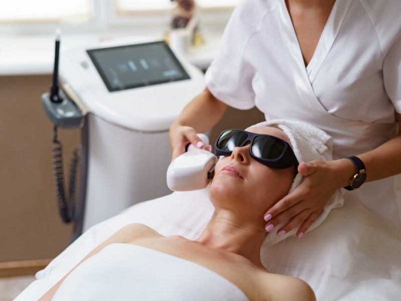 View of doctor cosmetologist doing anti aging procedure in cosmetology office. Satisfied woman in disposable hat lying on couch and relaxing. Working with Apparatus.